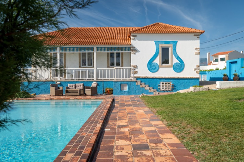A casa vista do outro lado da piscina, com o muro sobre o mar e o jardim florido