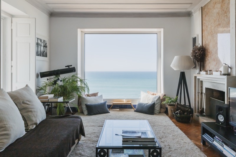 Sala de estar com sofá creme, lareira de pedra e janela panorâmica sobre o Atlântico ao entardecer